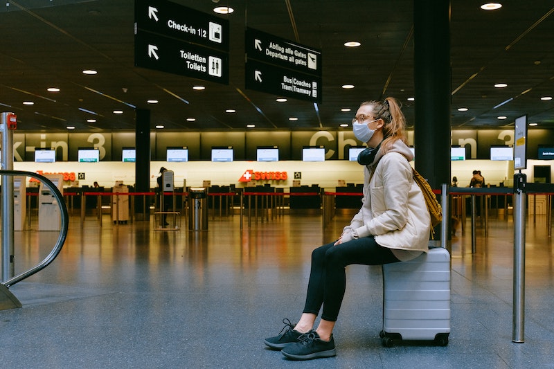 Woman wearing protective mask, seated at airport on her luggage